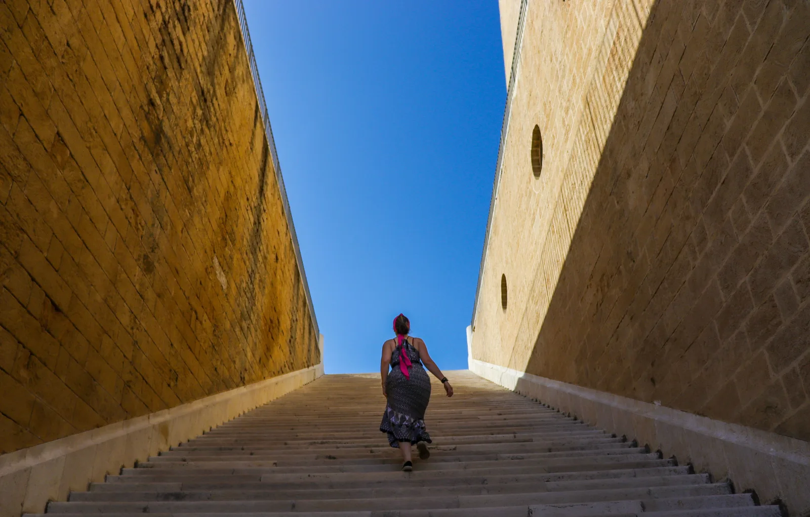 A woman walks up a broad staircase between tall stone walls.