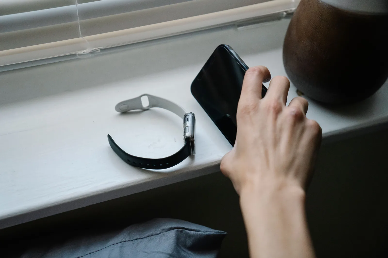 A hand reaches for a phone beside a smartwatch on a windowsill in the morning.