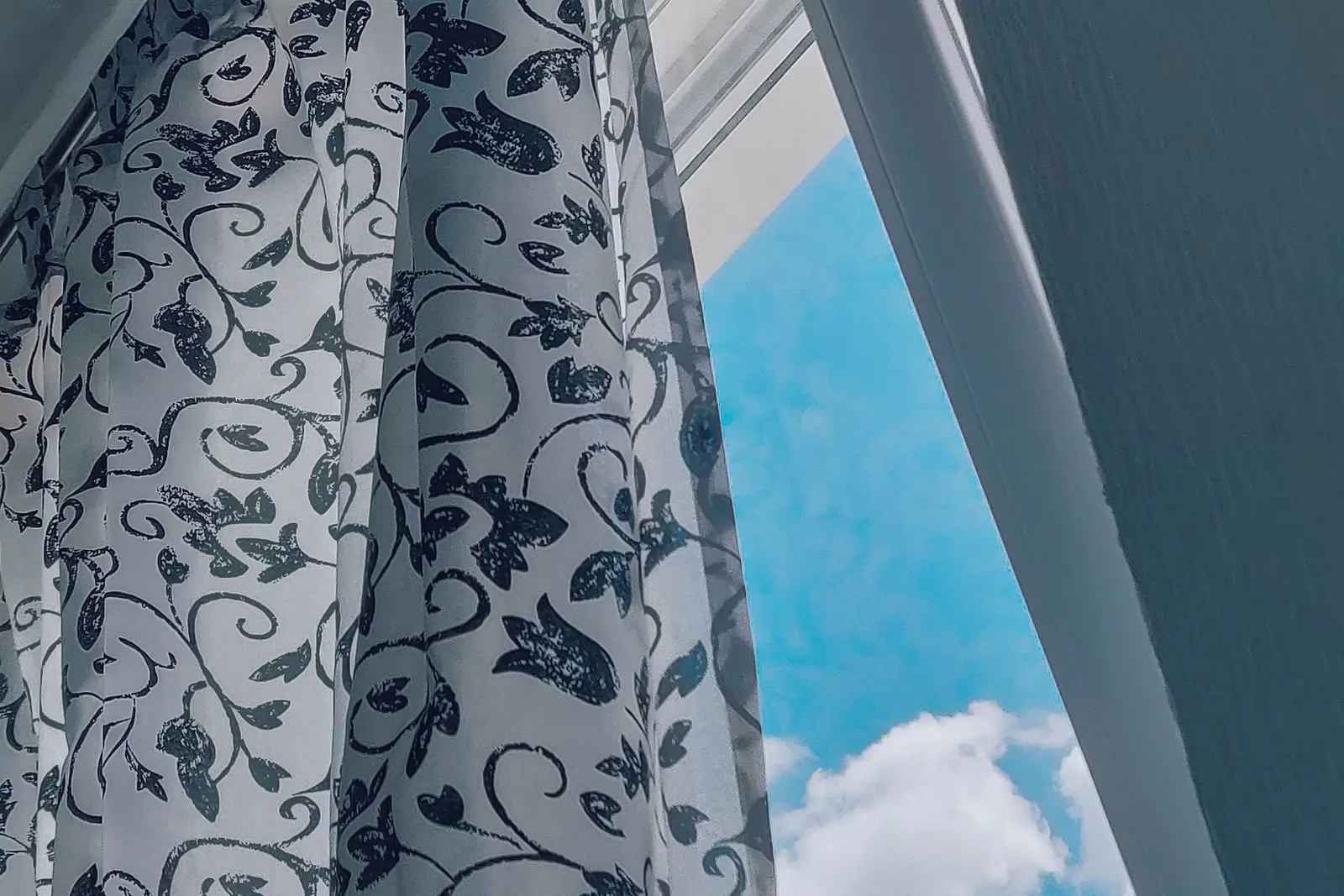 A patterned curtain beside an opened window with a blue summer sky.