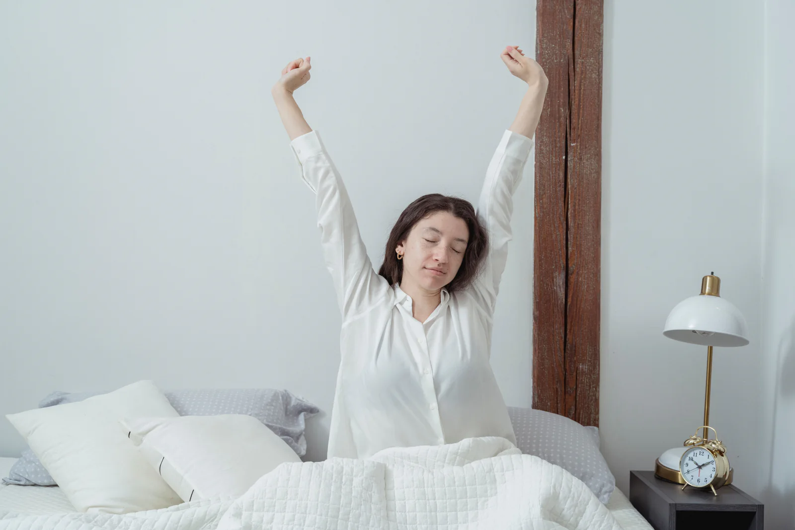 A woman stretches in bed in the morning beside an alarm clock.