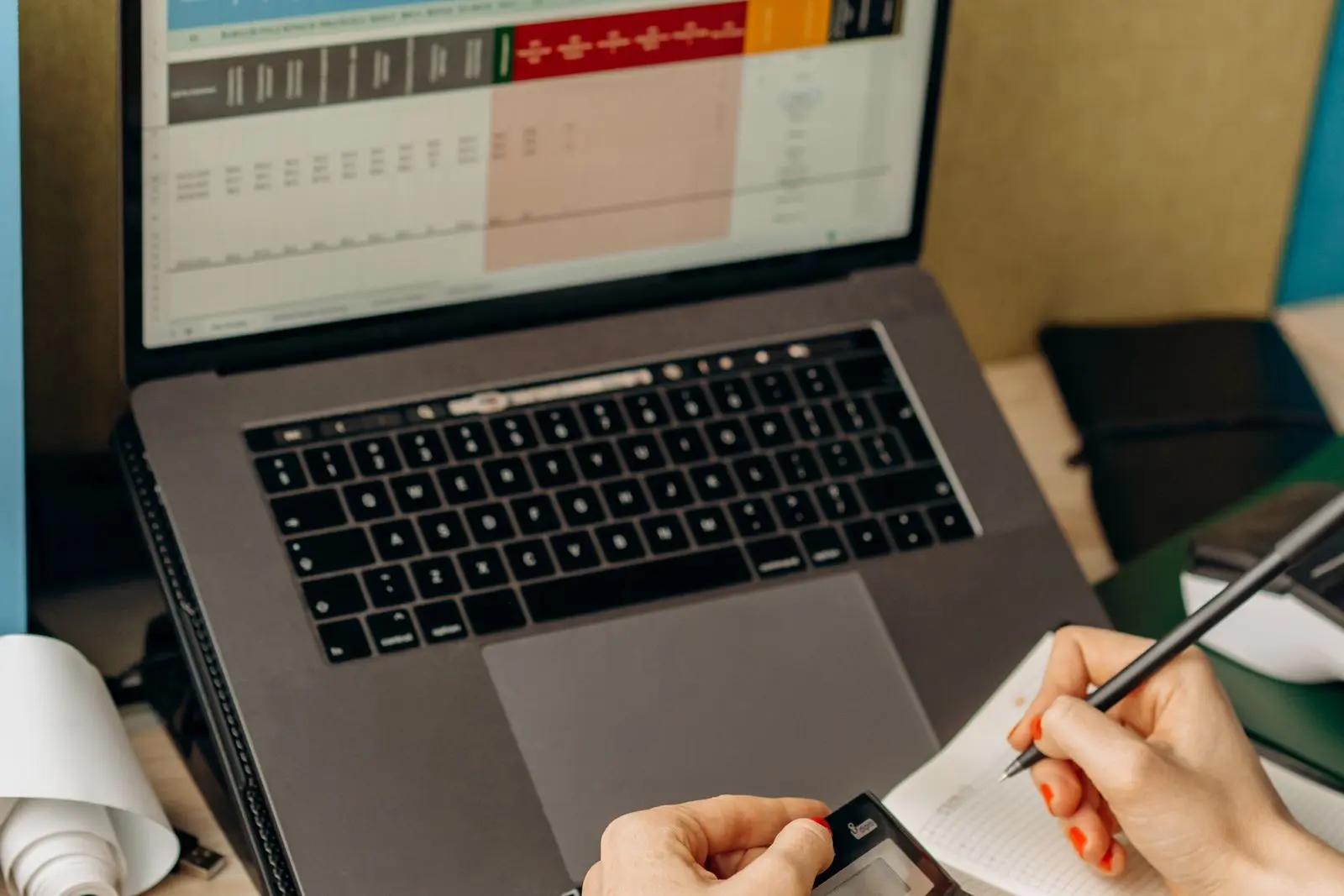 Hands taking notes next to a laptop with an open spreadsheet.